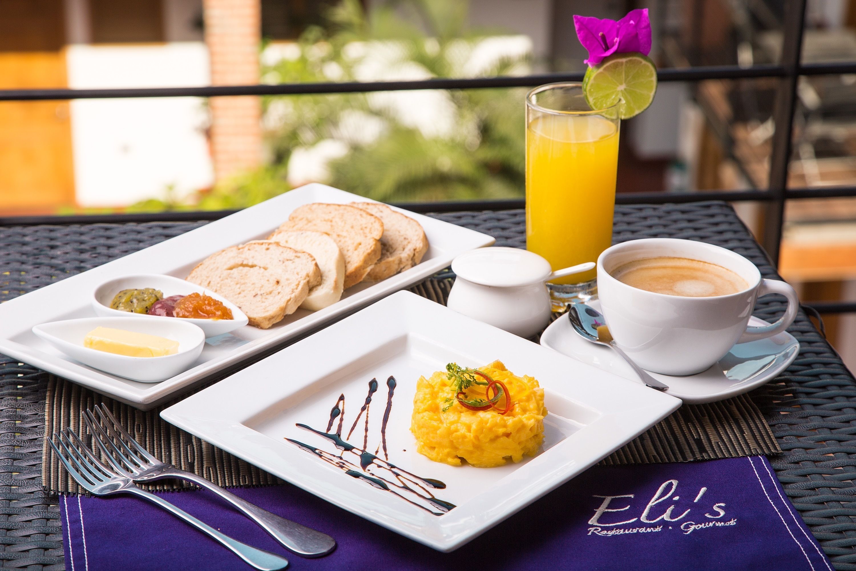Un desayuno servido con huevos revueltos, tostadas y mermelada, un vaso de jugo de naranja y una taza de café, todo sobre una mesa con un mantel individual que dice “Eli’s Restaurant & Gourmet”.