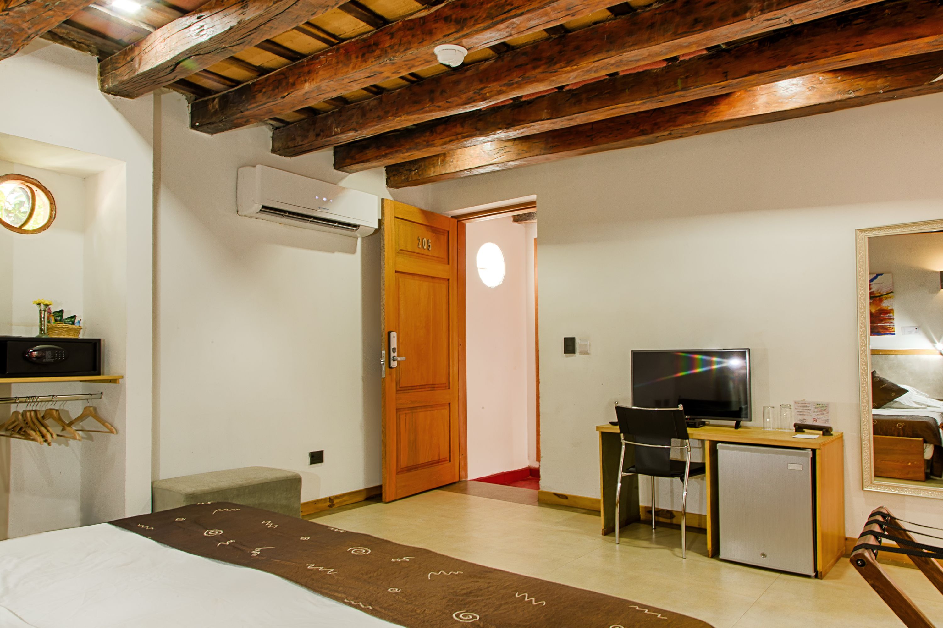 A hotel room with a wooden ceiling, a bed with a brown bedspread, a desk with a television, and a small refrigerator.