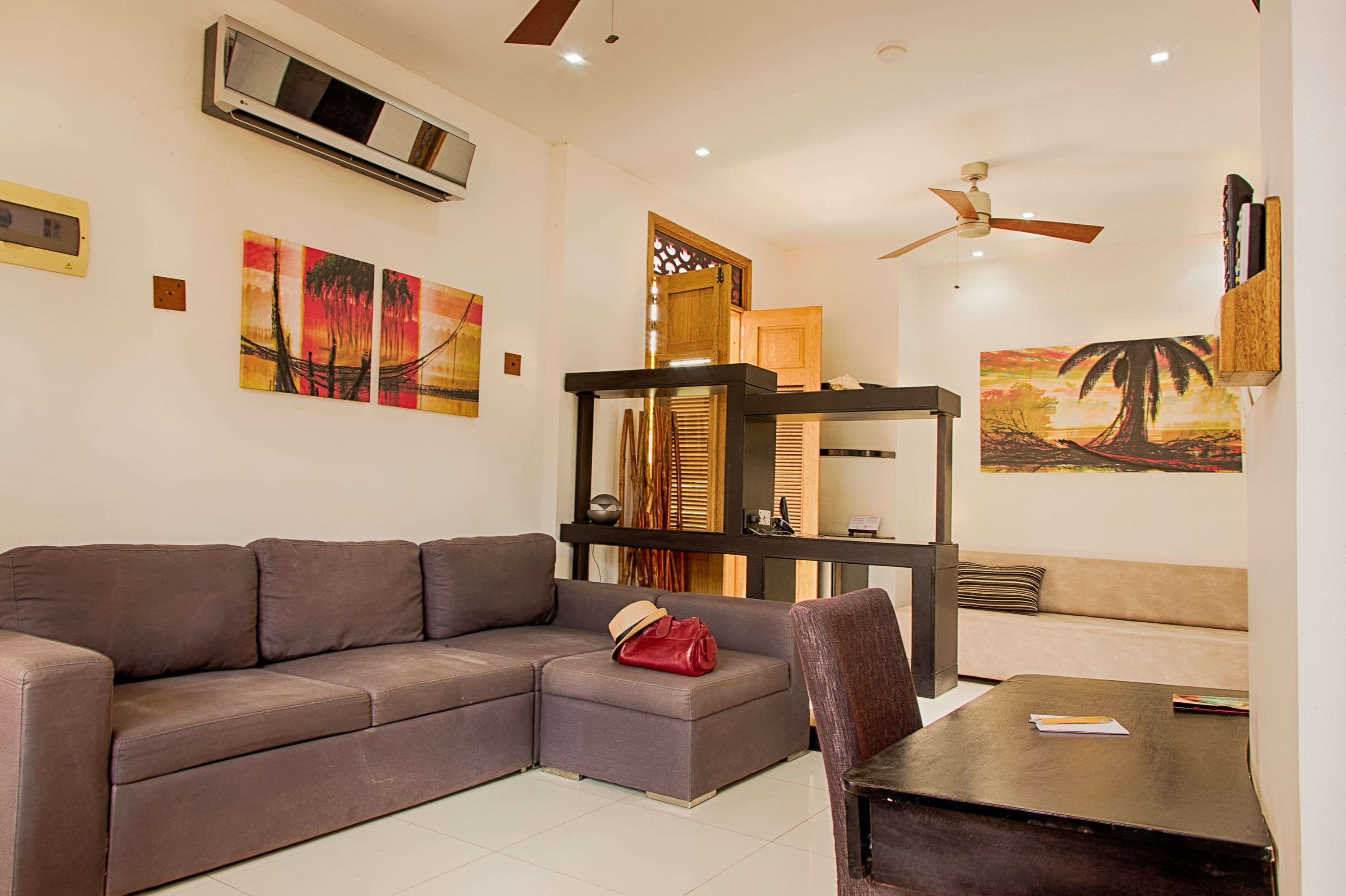 A living room with a sectional sofa, a small table, and a ceiling fan. There are paintings on the walls and a wooden cabinet.