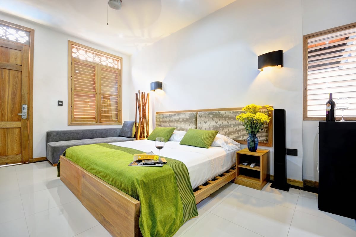 A modern bedroom with a wooden bed, green bedding, a bedside table with a vase of flowers, and a nightstand with a wine glass.