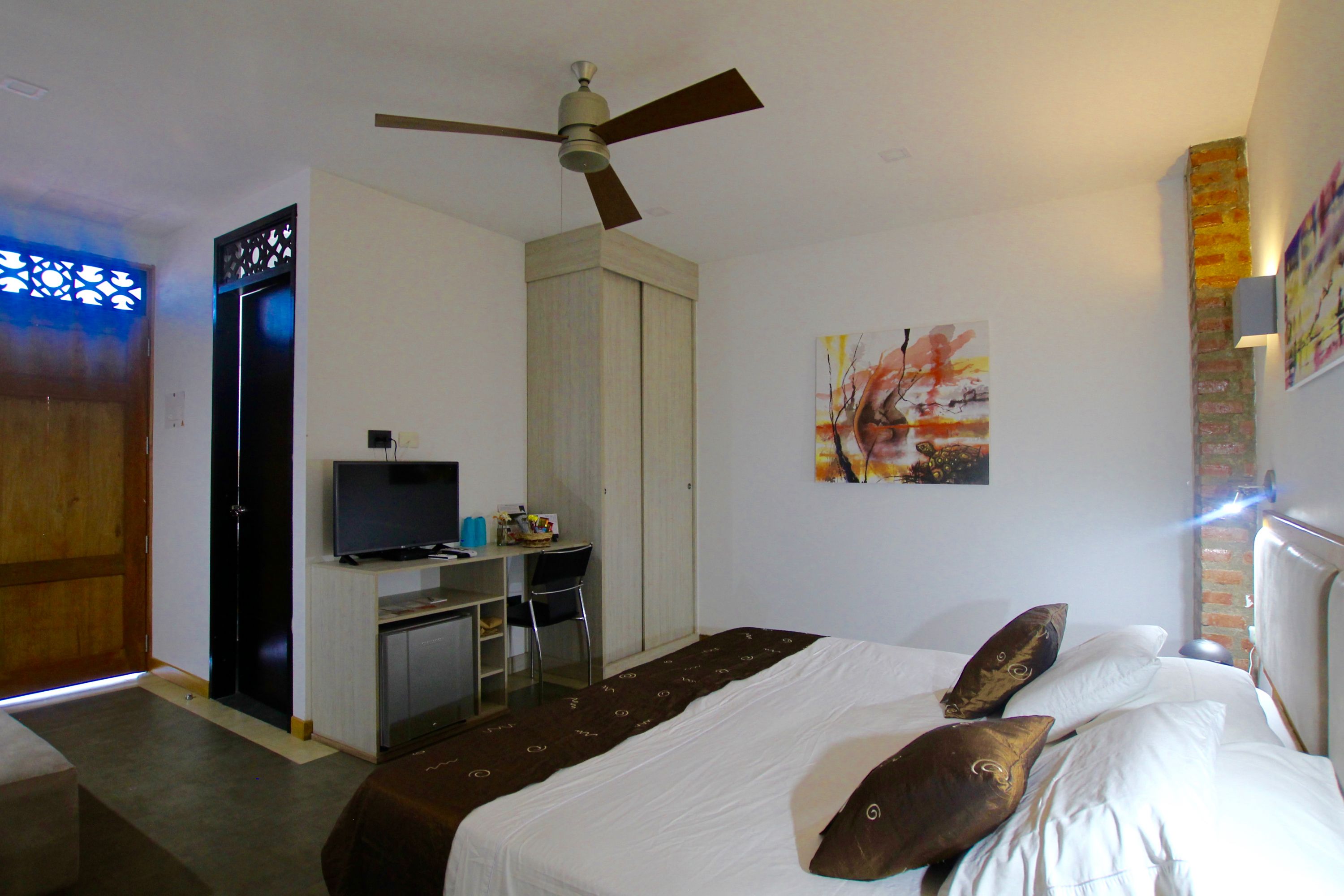 Una habitación de hotel con cama, escritorio con televisor, ventilador de techo y cuadros en la pared.