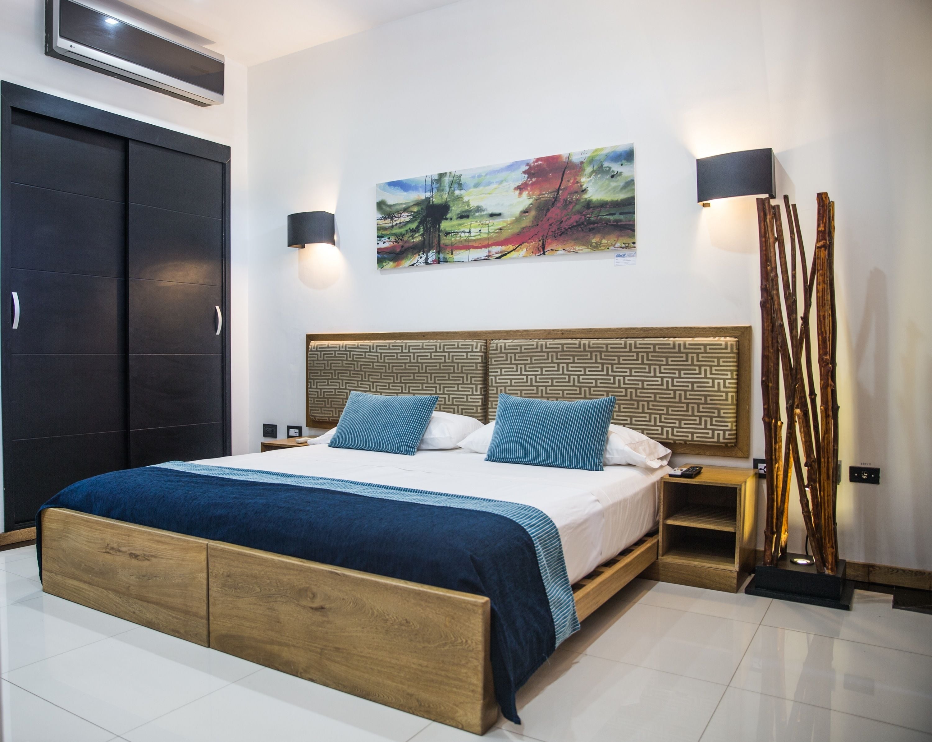A bedroom with a wooden bed, a nightstand, a wall-mounted air conditioner, a painting, and a tall floor lamp.