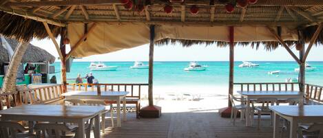 A beachside restaurant with wooden tables and chairs, a thatched roof, and a view of the sea with boats.