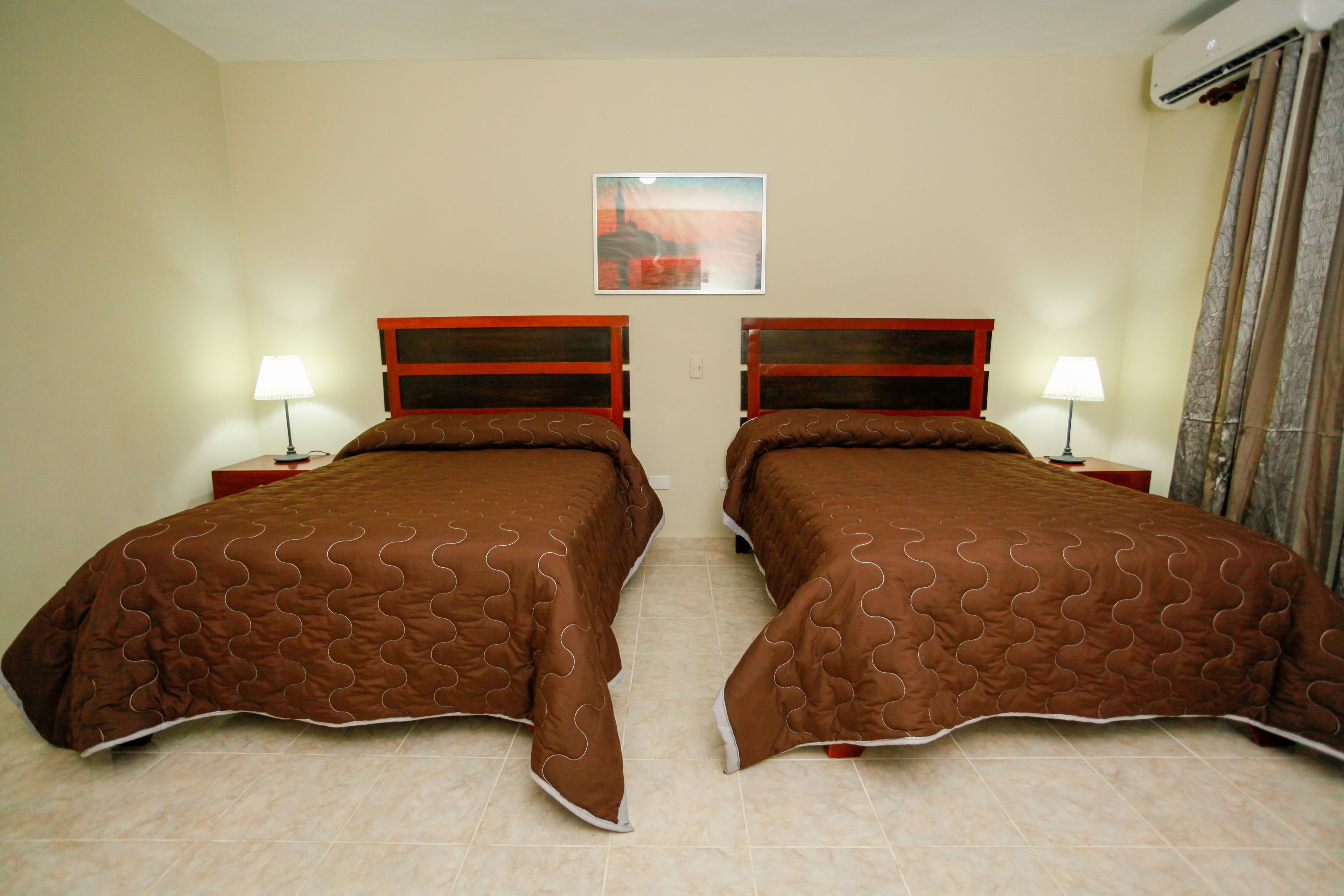A hotel room with two beds, each with a wooden headboard and a brown quilted bedspread. There is a single wall-mounted air conditioning unit and a framed picture above the beds.