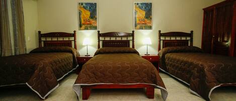 Habitación de hotel con tres camas individuales, cada una con cabecero de madera y colcha marrón acolchada. Dos lámparas de pared a cada lado de la cama central. El piso es de baldosas y las paredes son color beige.