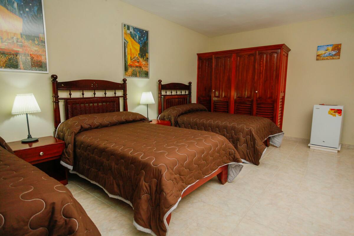 A hotel room with two beds, wooden headboards, a wardrobe, a refrigerator, and artwork on the walls.