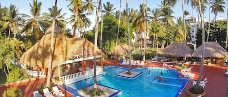 Zona de piscina en un resort con varias piscinas, sillas de descanso y estructuras de techo de paja.