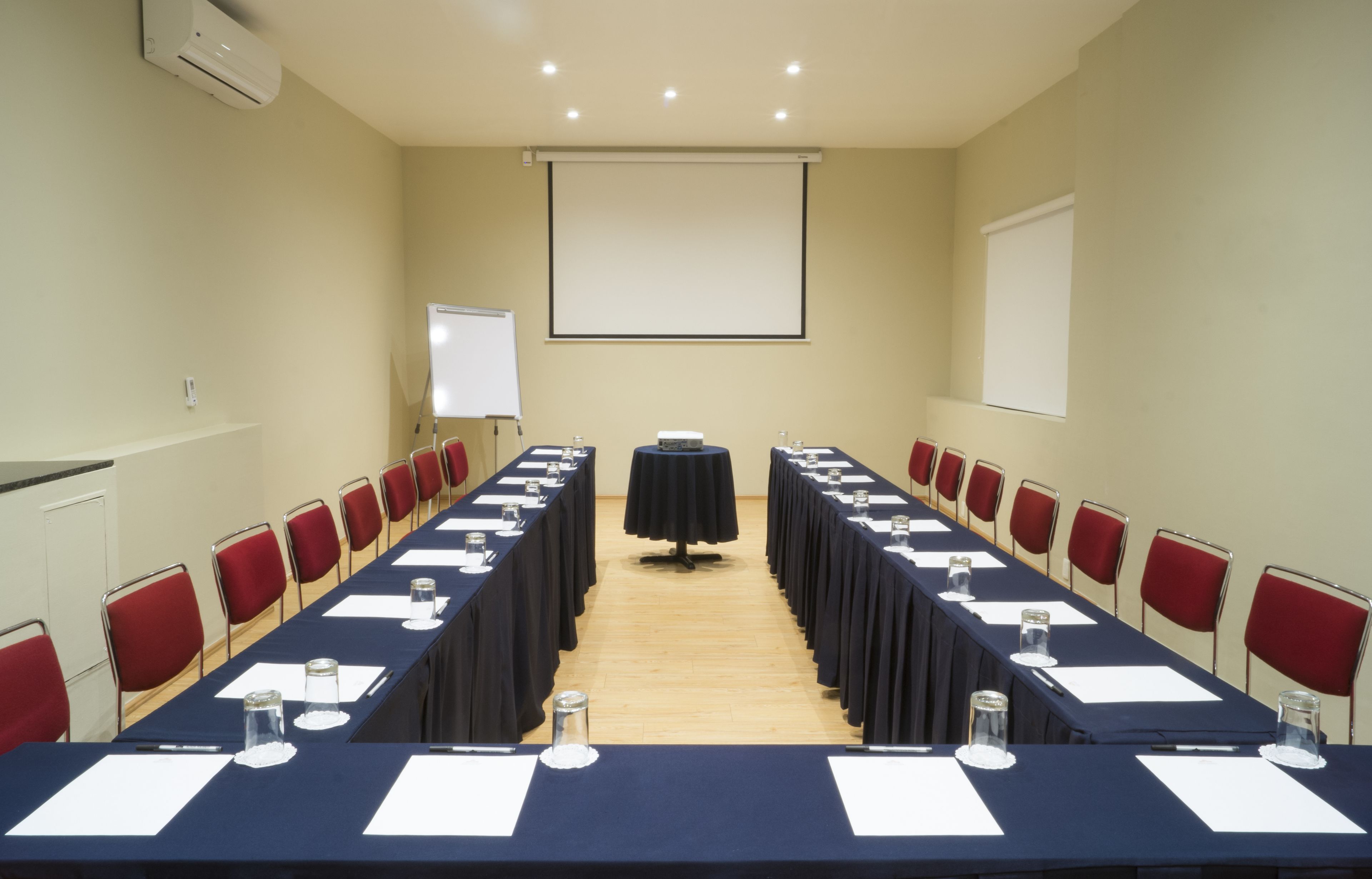 Un salón de conferencias con una larga mesa, sillas rojas, una pantalla de proyector y una pizarra.