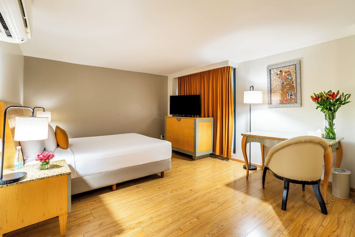 A hotel room with a bed, a desk, a chair, a TV, a lamp, and a vase with flowers.