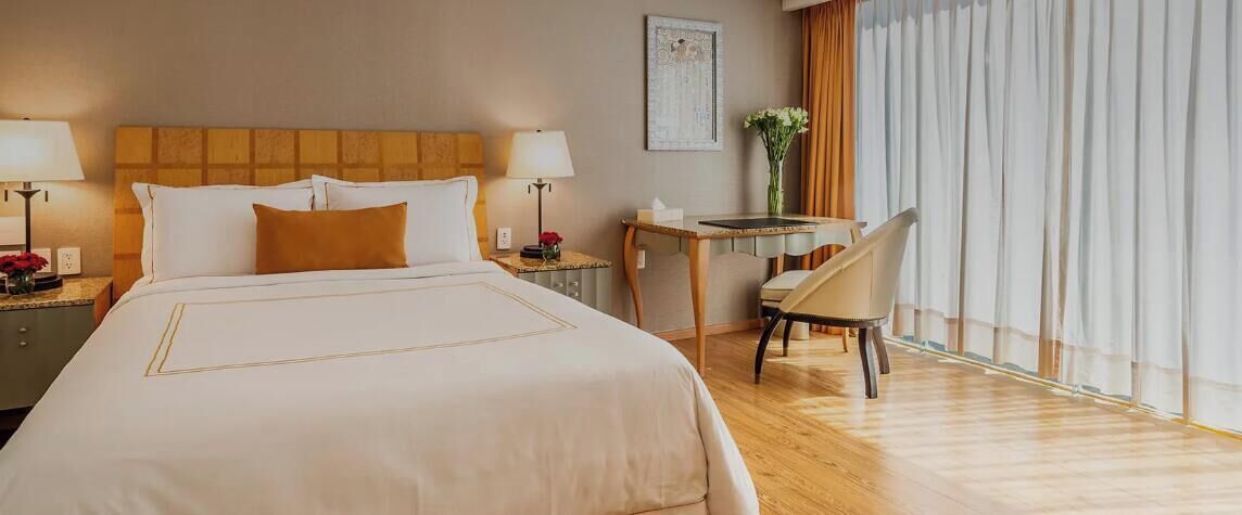 A hotel room with a large bed, bedside tables, a desk, a chair, and a window with curtains.