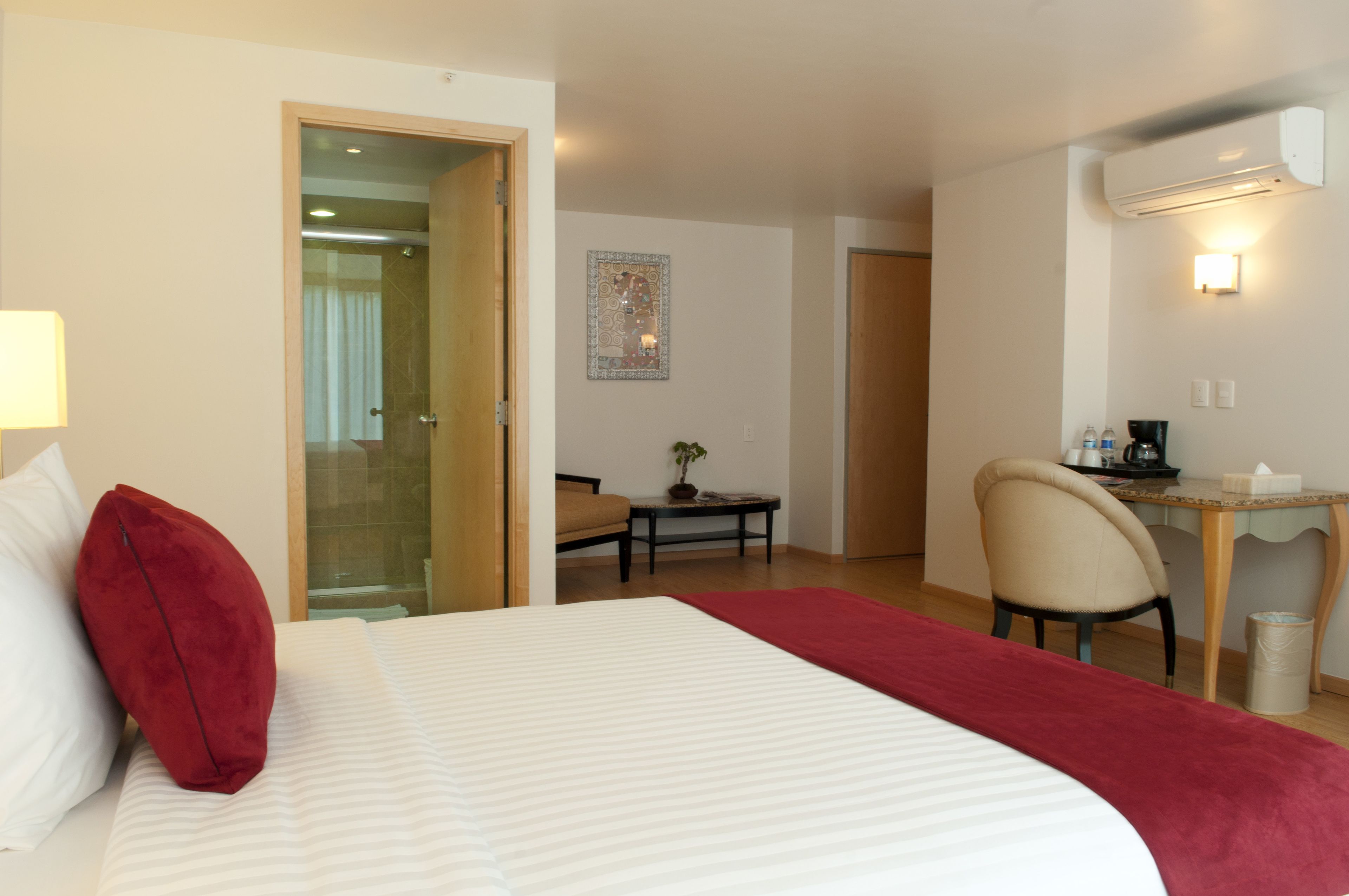 A hotel room with a bed, a desk, a chair, a small table with a plant, and an air conditioning unit.