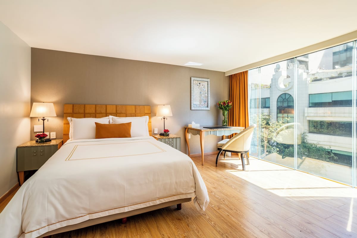 A hotel room with a large bed, two bedside tables with lamps, a desk with a chair, and a large window offering a view of the city.