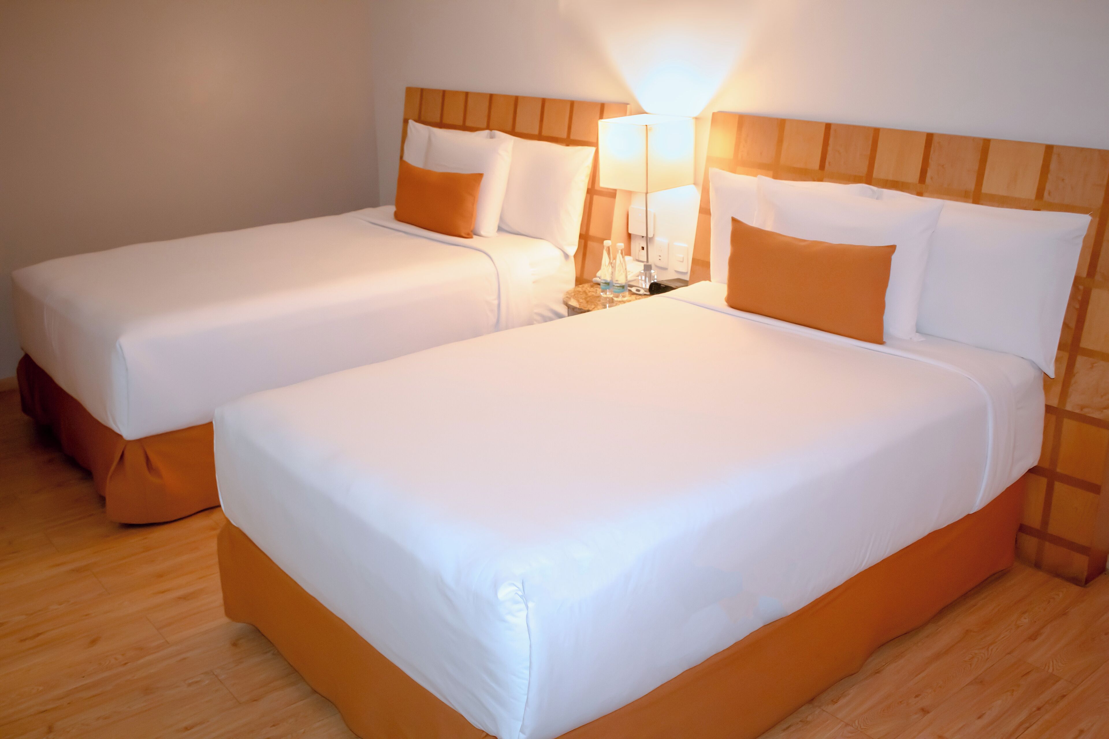 A hotel room with two beds, each with white bedding and orange accents, a wooden headboard, and a nightstand with a lamp.