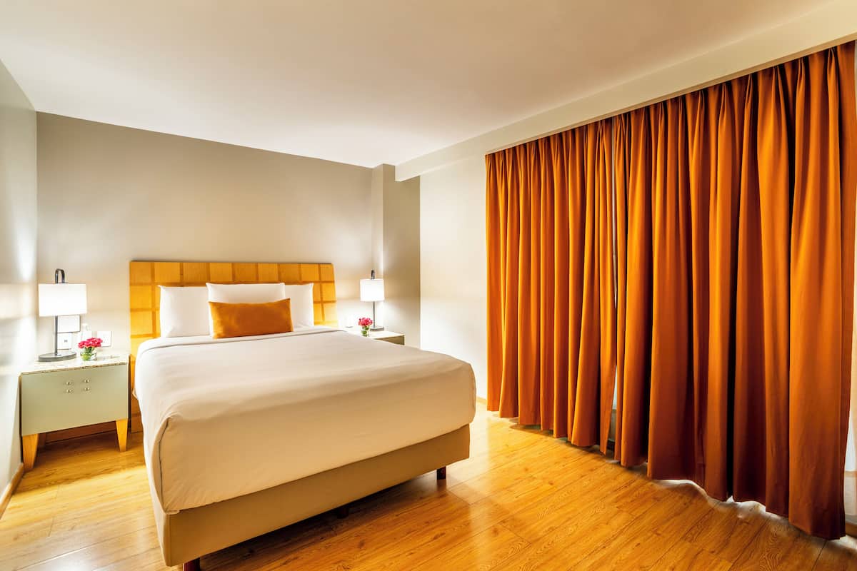 A hotel room with a large bed, wooden flooring, a nightstand with a lamp, and a window with orange curtains.