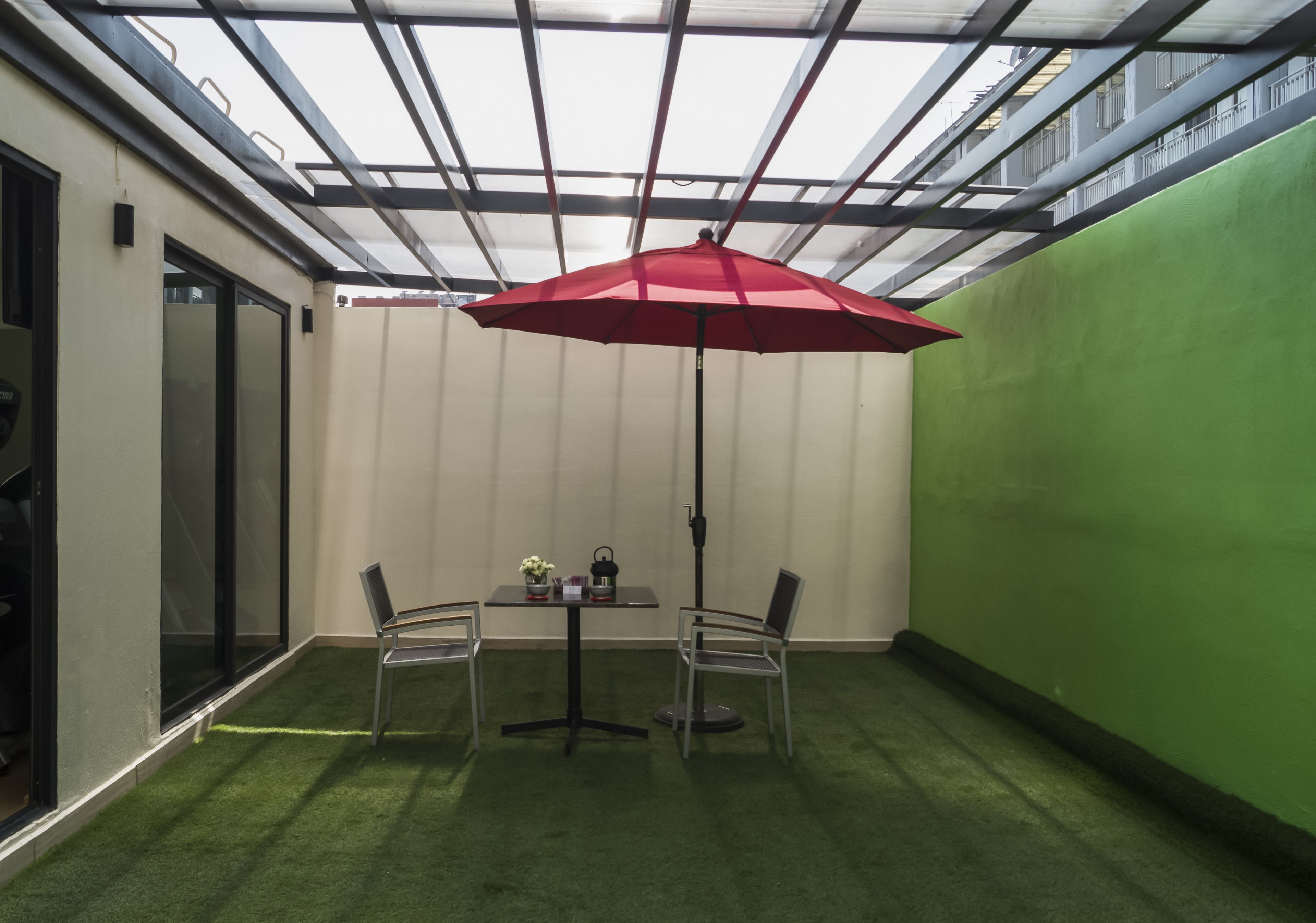 Zona de pati petita i tancada amb una taula, dues cadires, un parasol vermell i una paret verda.