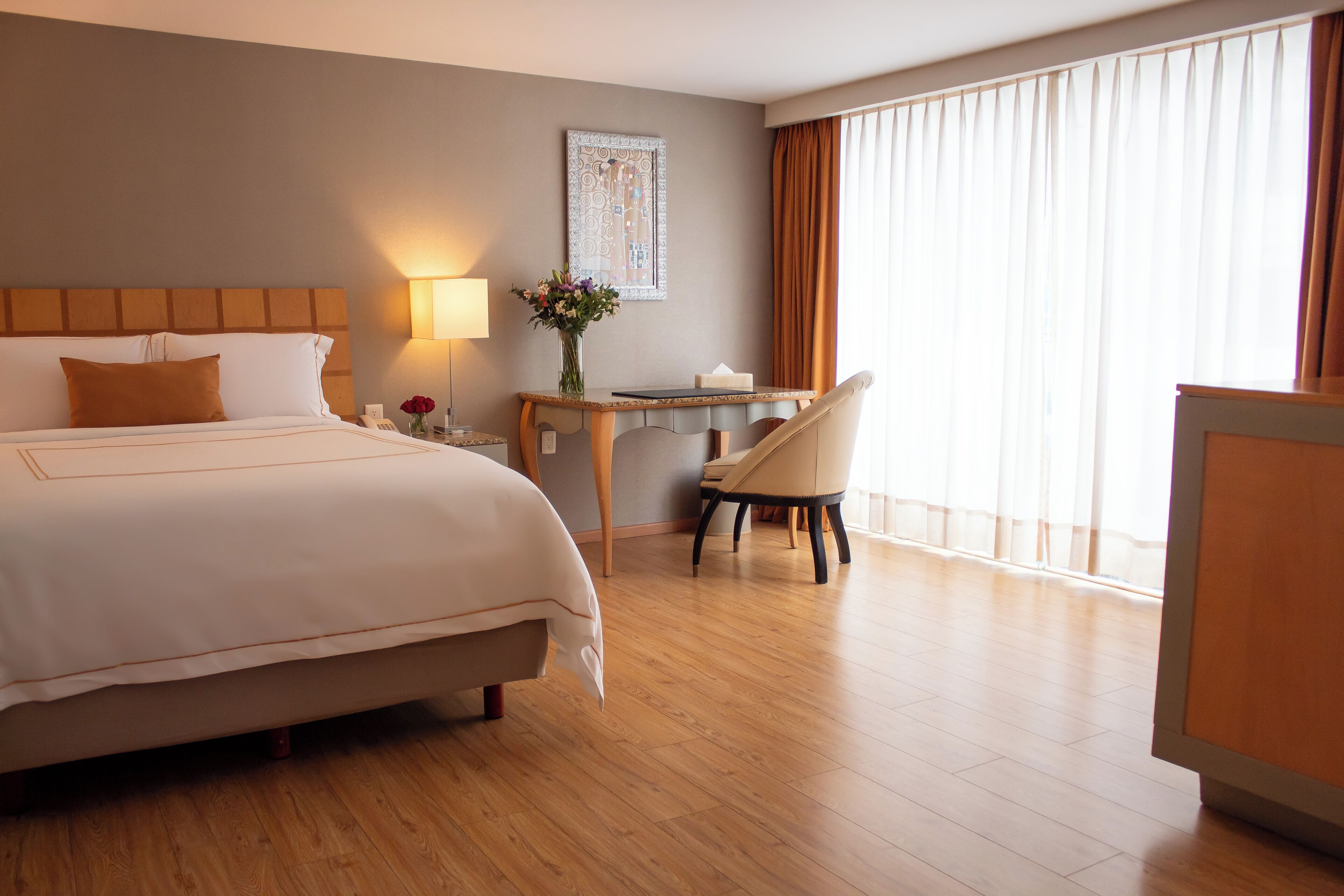 A hotel room with a large bed, a desk with a chair, a lamp, and a vase with flowers.