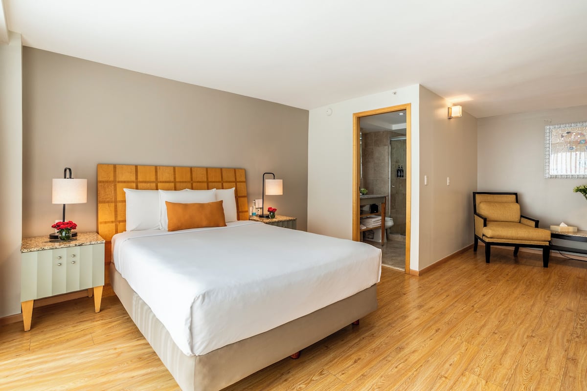 A hotel room with a large bed, two bedside tables with lamps, a chair, and a door leading to a bathroom.