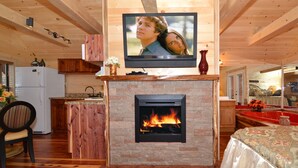 Interior acogedor de una cabaña con chimenea, pisos de madera, un televisor montado y una zona de cocina.