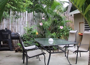 A glass-top table with four chairs, a barbecue grill, and a bamboo fence in a garden setting.