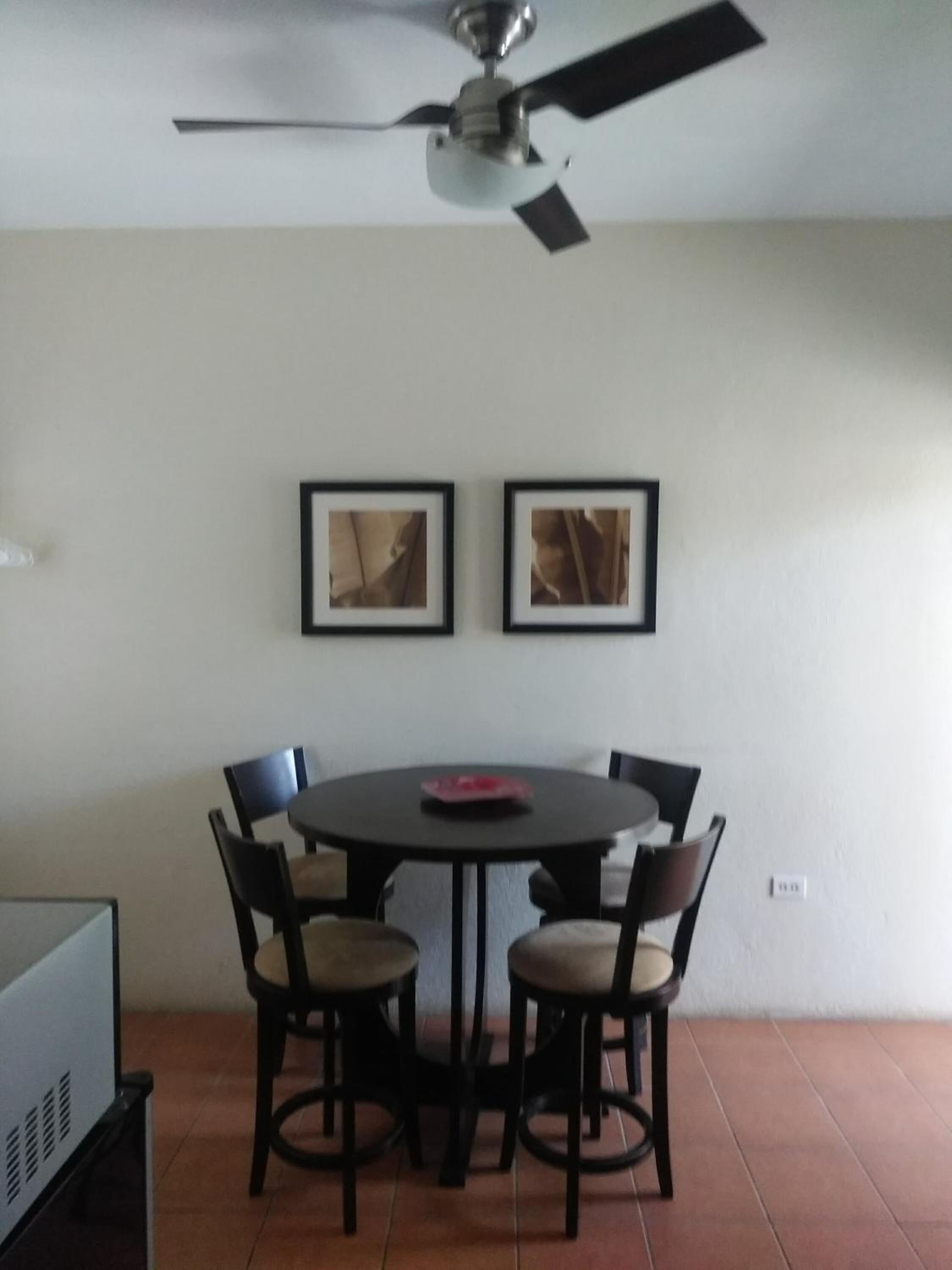 A dining area with a round table and six chairs, a ceiling fan, and two framed pictures on the wall.