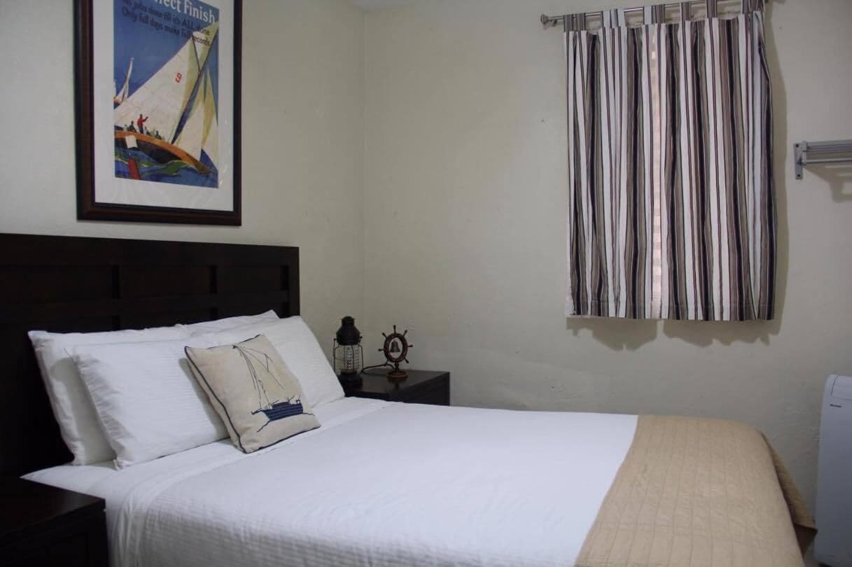 A neatly made bed with white bedding and a beige throw blanket, a framed poster on the wall, a striped curtain, and a small table with a lantern and a ship's wheel.