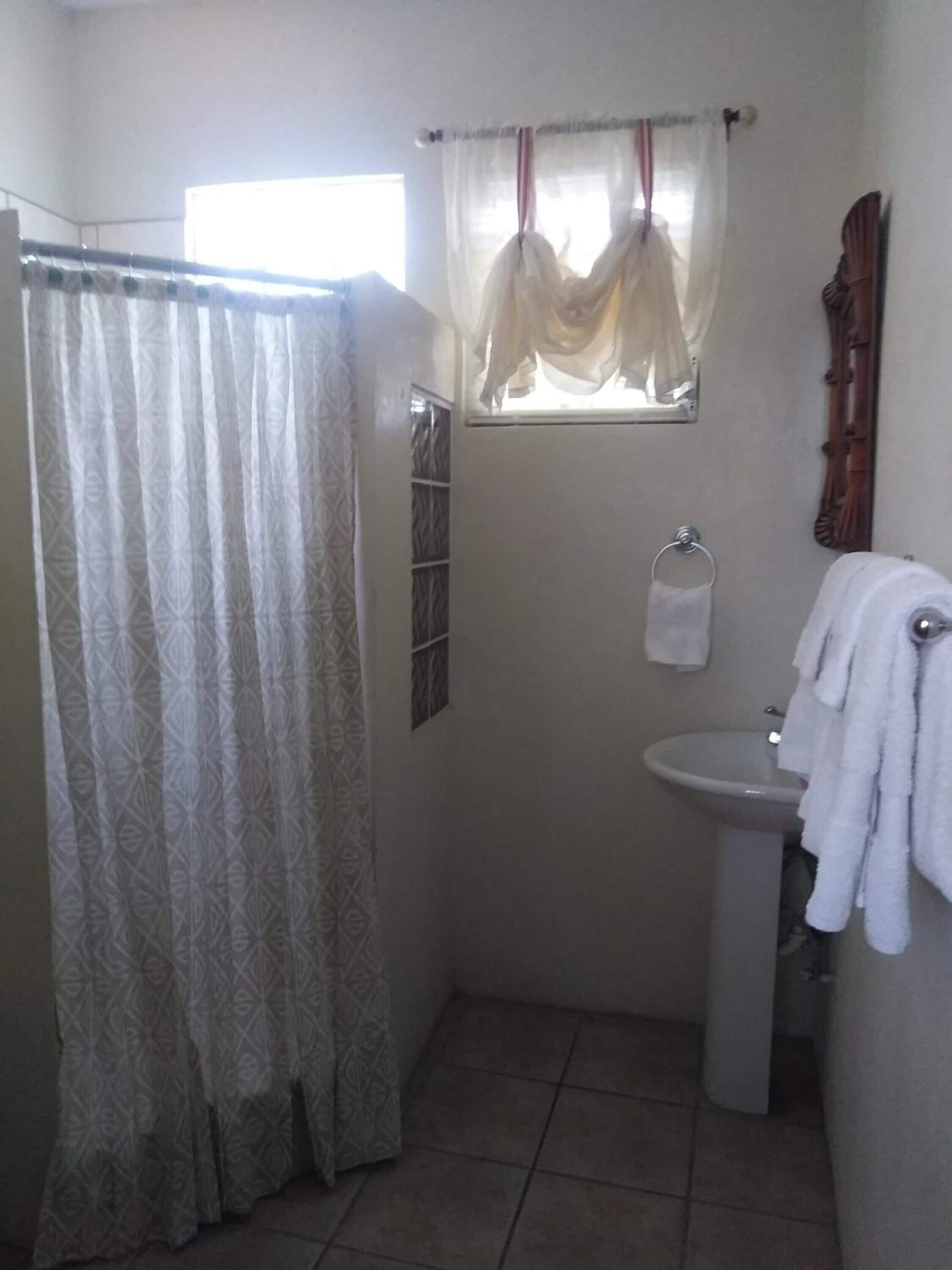 Un baño con cortina de ducha, una ventana con cortinas, un lavamanos y un perchero para toallas.