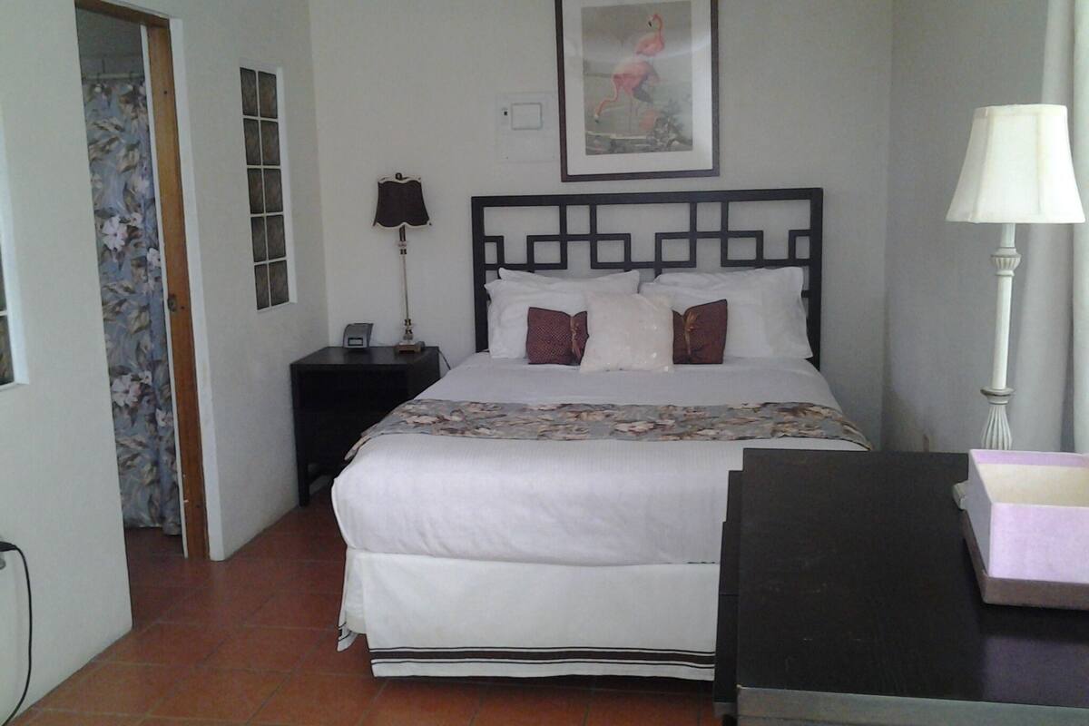 A bedroom with a bed, a nightstand, a lamp, a framed picture on the wall, and a door with floral wallpaper.