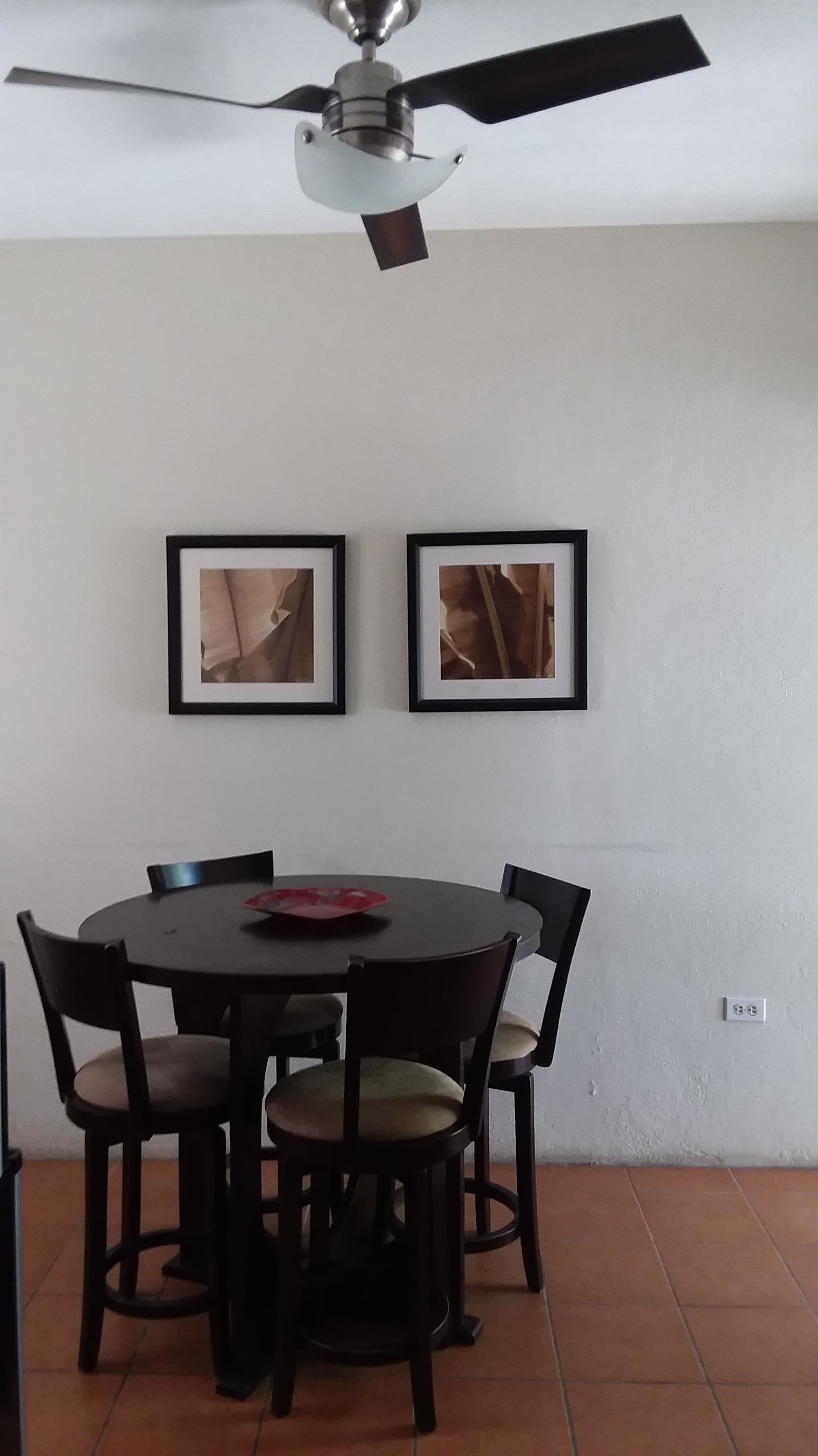 A dining area with a round table, six chairs, two framed pictures on the wall, and a ceiling fan.