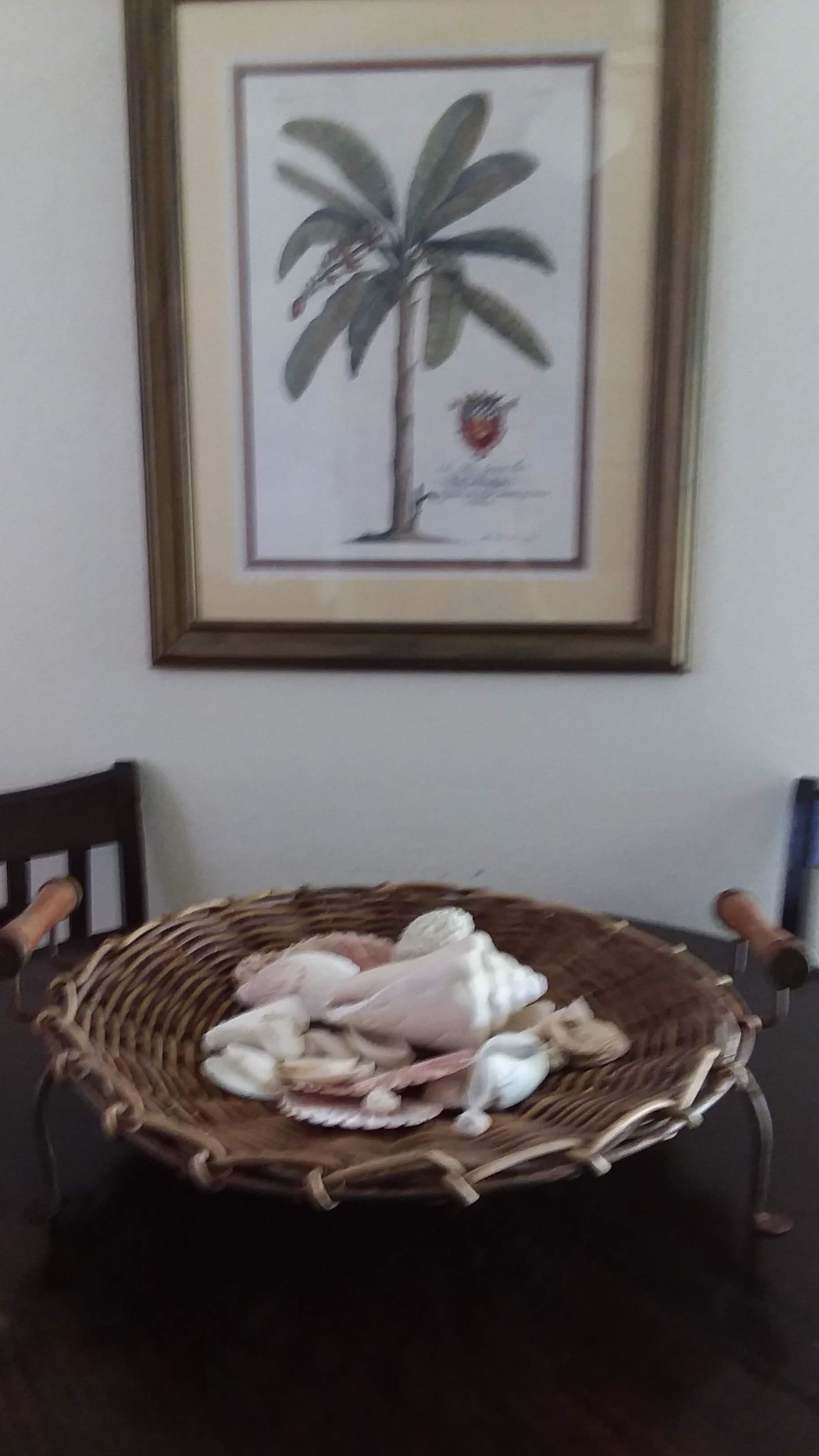 Une œuvre d’art encadrée représentant un palmier sur un mur, un panier en osier contenant un objet blanc sur une table, et deux chaises.