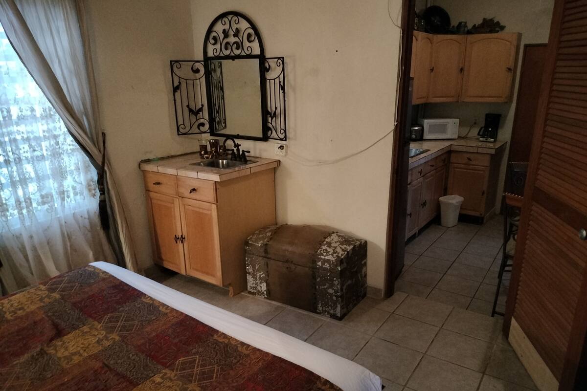 A bedroom with a bed, a wooden cabinet, a small stool, a mirror, and a kitchenette in the background.