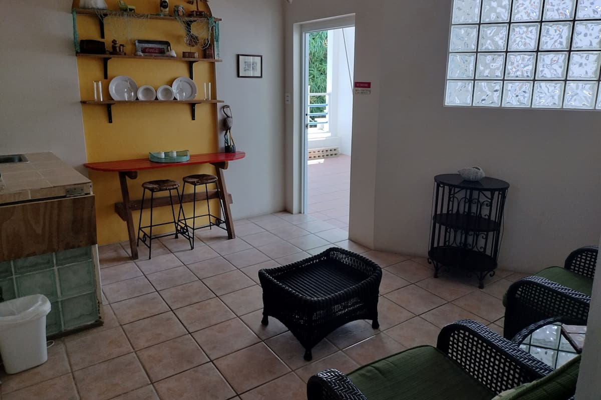 A room with a tiled floor, a red table with stools, a wicker bench, and a black chair. There is a shelf with dishes and a framed picture.