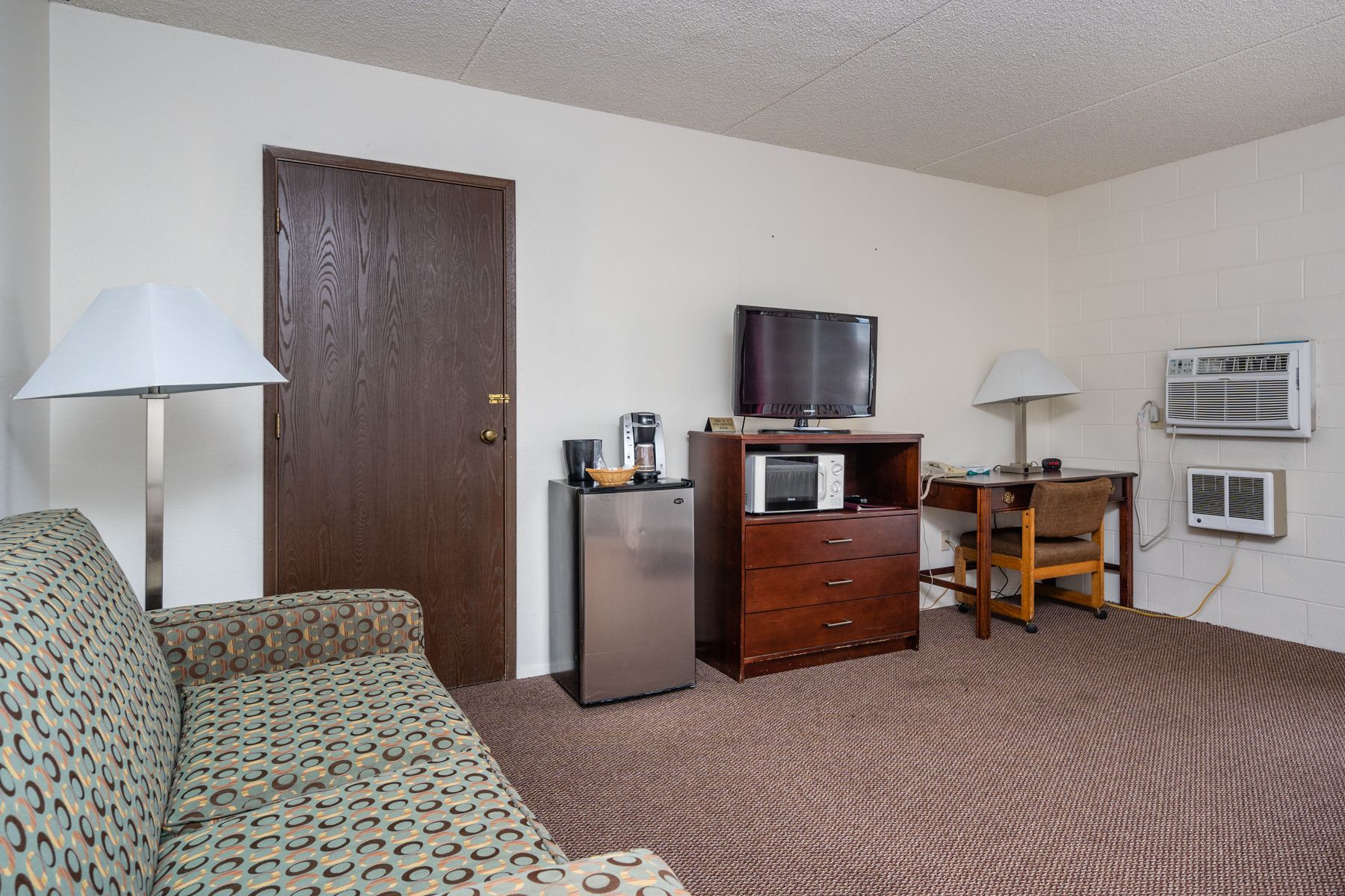 A compact hotel room with a sofa, a desk with a computer, a television, a refrigerator, and an air conditioning unit.