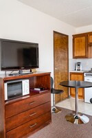 Quarto de hotel compacto com cômoda de madeira, micro-ondas, TV Samsung, mesa pequena para refeições e cozinha americana com cafeteira.