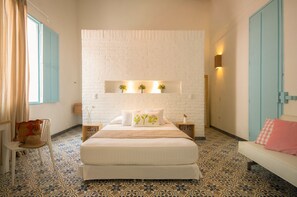 Una camera da letto con una parete in mattoni bianchi, un letto con biancheria bianca, una sedia blu, un tavolino con un vaso e una porta blu.