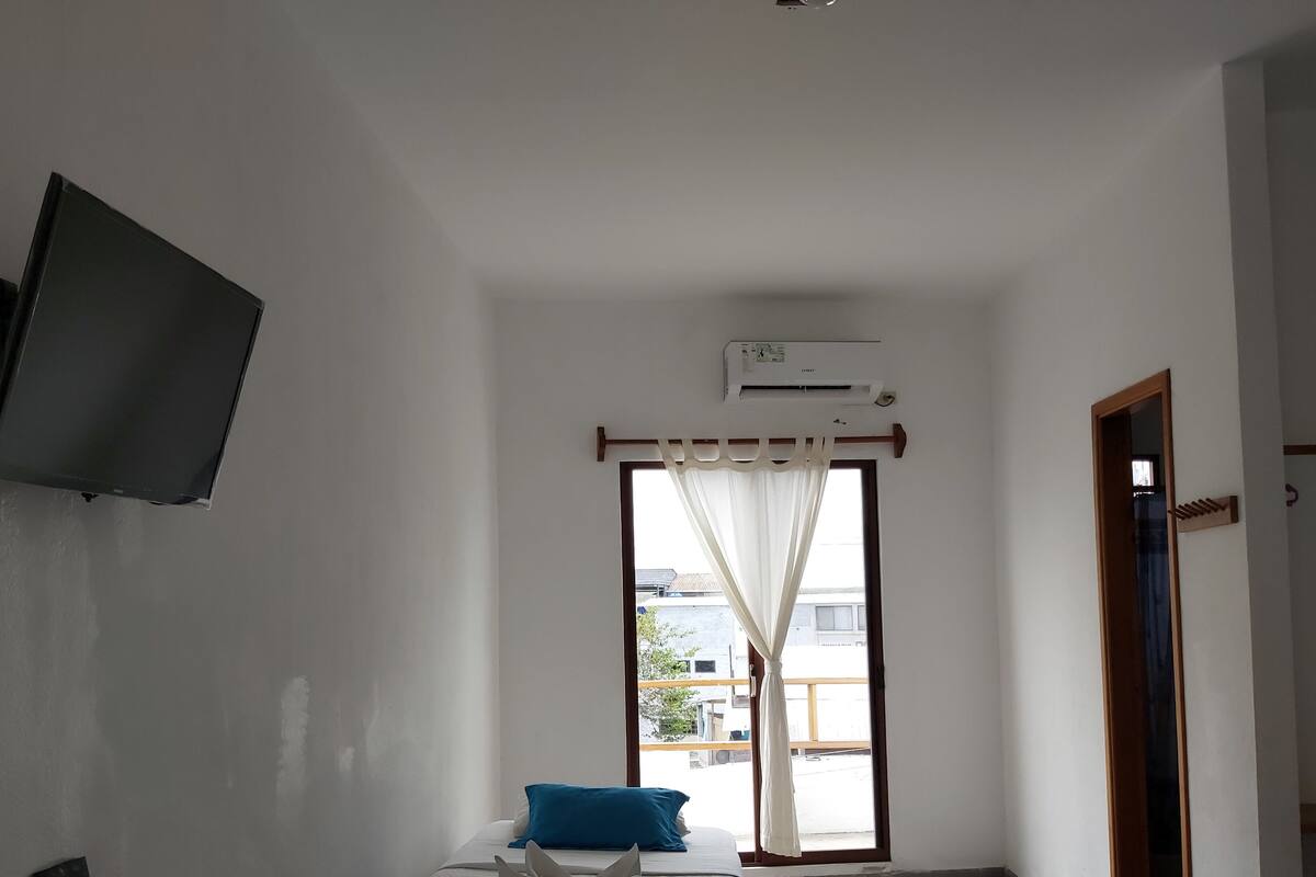 A room with a bed, a TV mounted on the wall, an air conditioning unit, and a window with curtains.