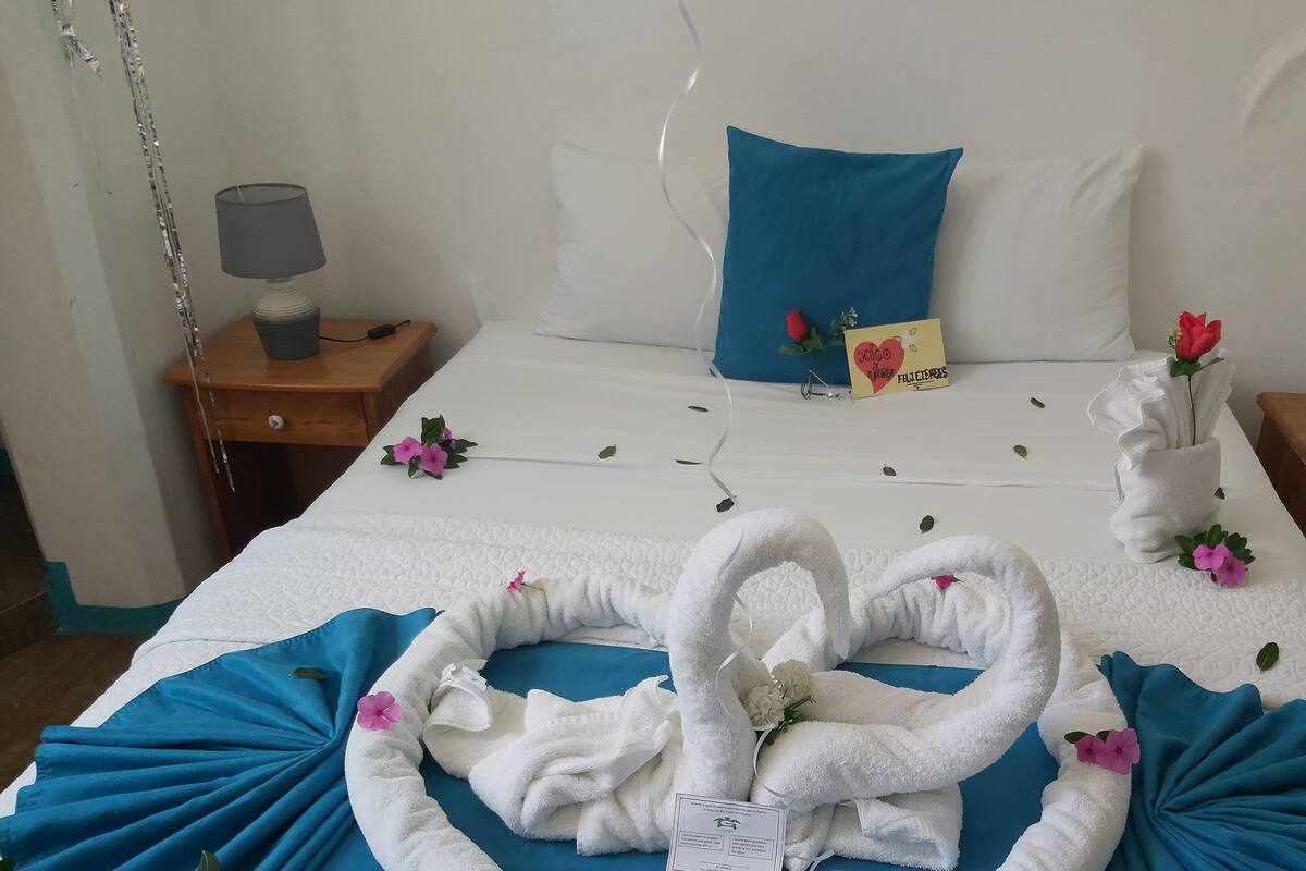 A neatly made bed with a blue pillow, a white pillow, and a card on the bed. There are towels arranged on the bed, a lamp on a bedside table, and a remote control on another bedside table.