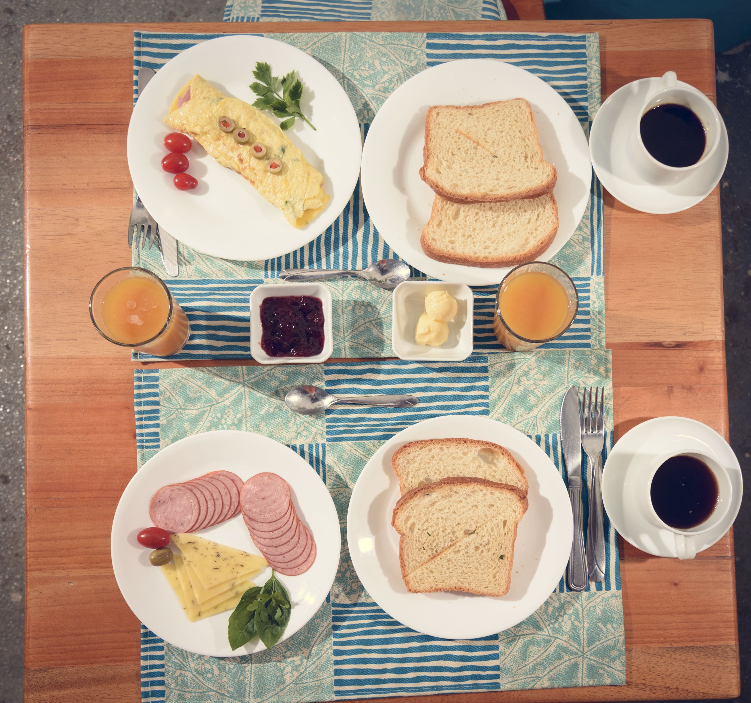 Una mesa de desayuno con platos de huevos, tostadas, jamón y queso, junto con tazas de café y jugo.