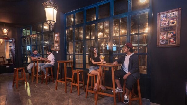 A bar area with wooden stools and tables, large windows with blue frames, and wall-mounted light fixtures. - Galapagos Native (Puerto Ayora)