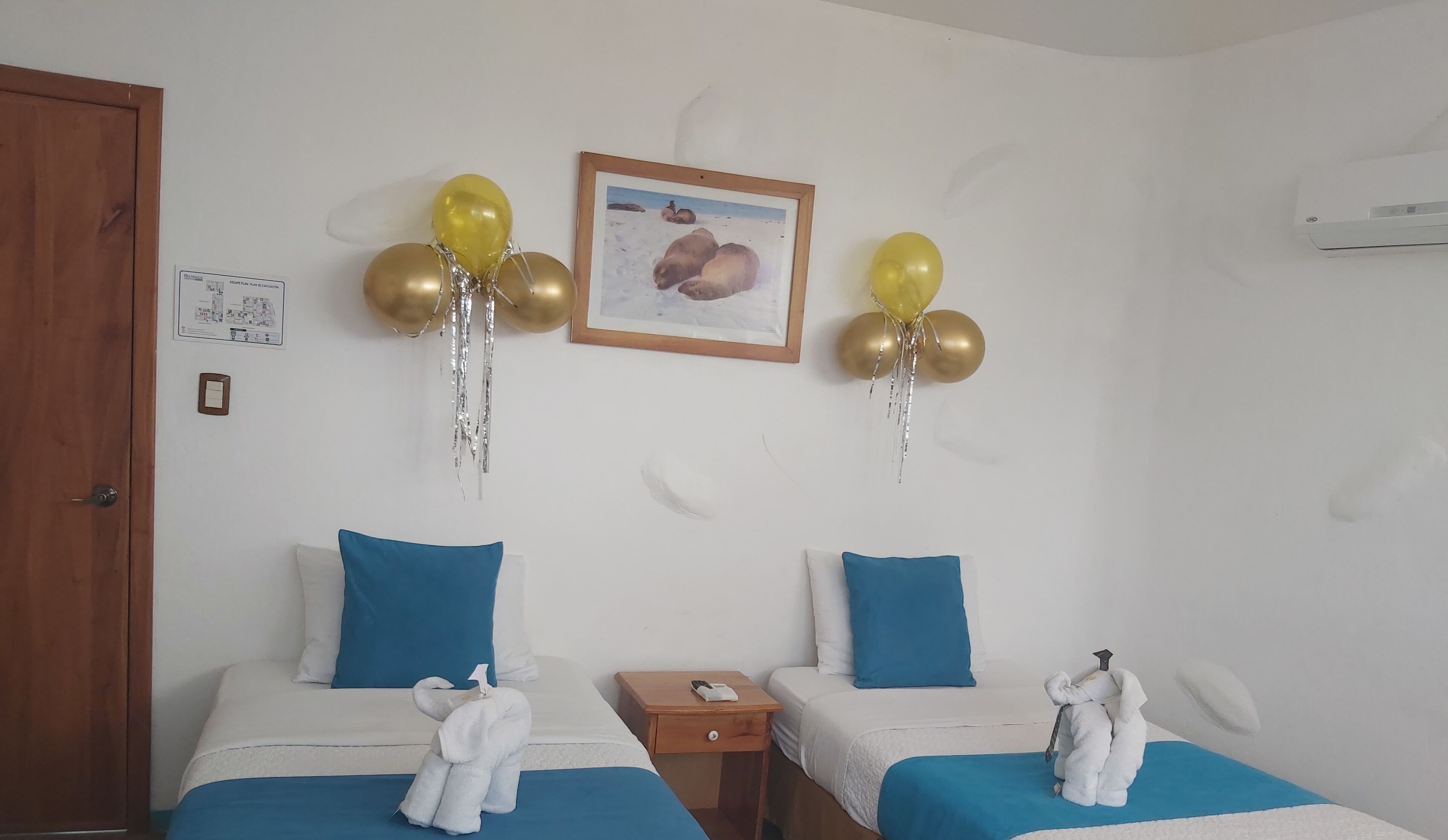 Een hotelkamer met twee bedden, elk met een blauw kussen en een witte deken. Er hangen gouden en zilveren ballonnen aan het plafond, en een lijst met een foto hangt aan de muur.