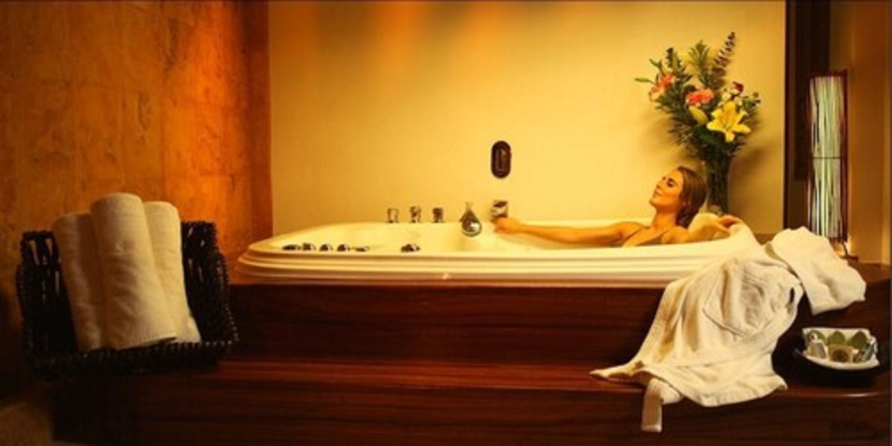 Una persona relajándose en una bañera, con una toalla, un jarrón de flores y una botella al lado.
