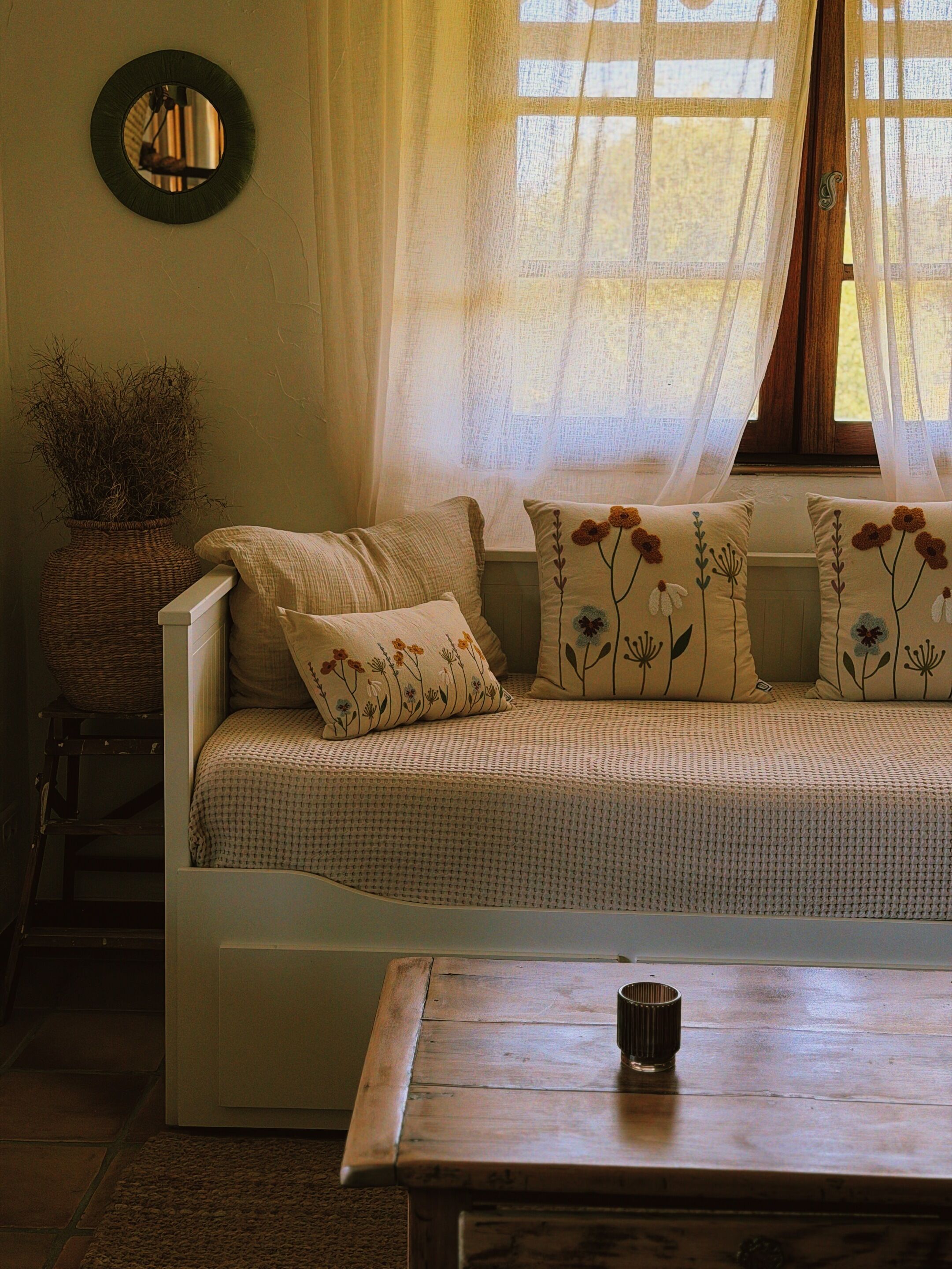 Una habitación acogedora con un sofá, una mesita, un jarrón con plantas secas y una ventana con cortinas translúcidas.