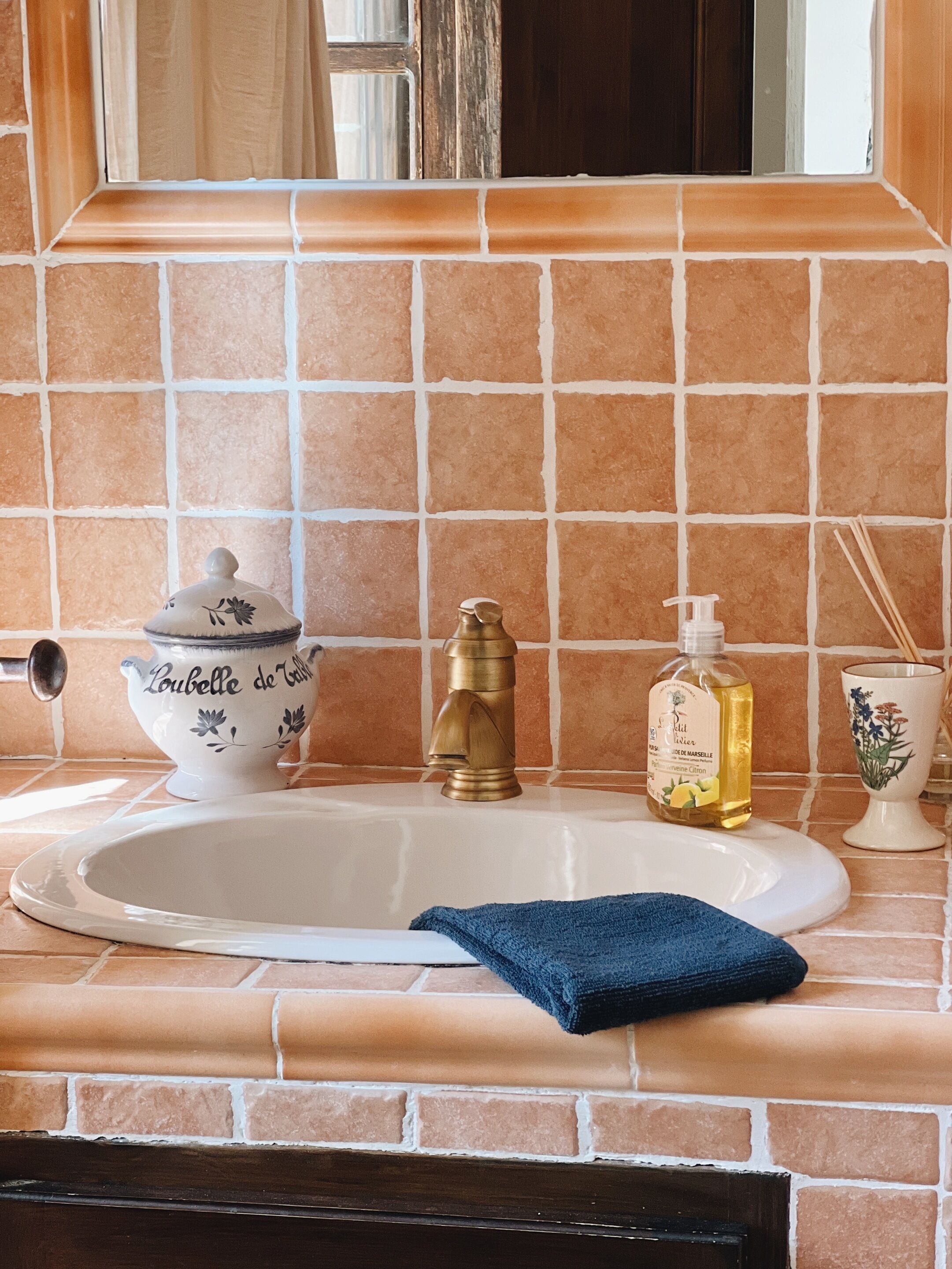 Un baño con un lavamanos blanco, un grifo dorado, un cuenco decorativo, una botella de jabón y una toalla azul sobre el mostrador.