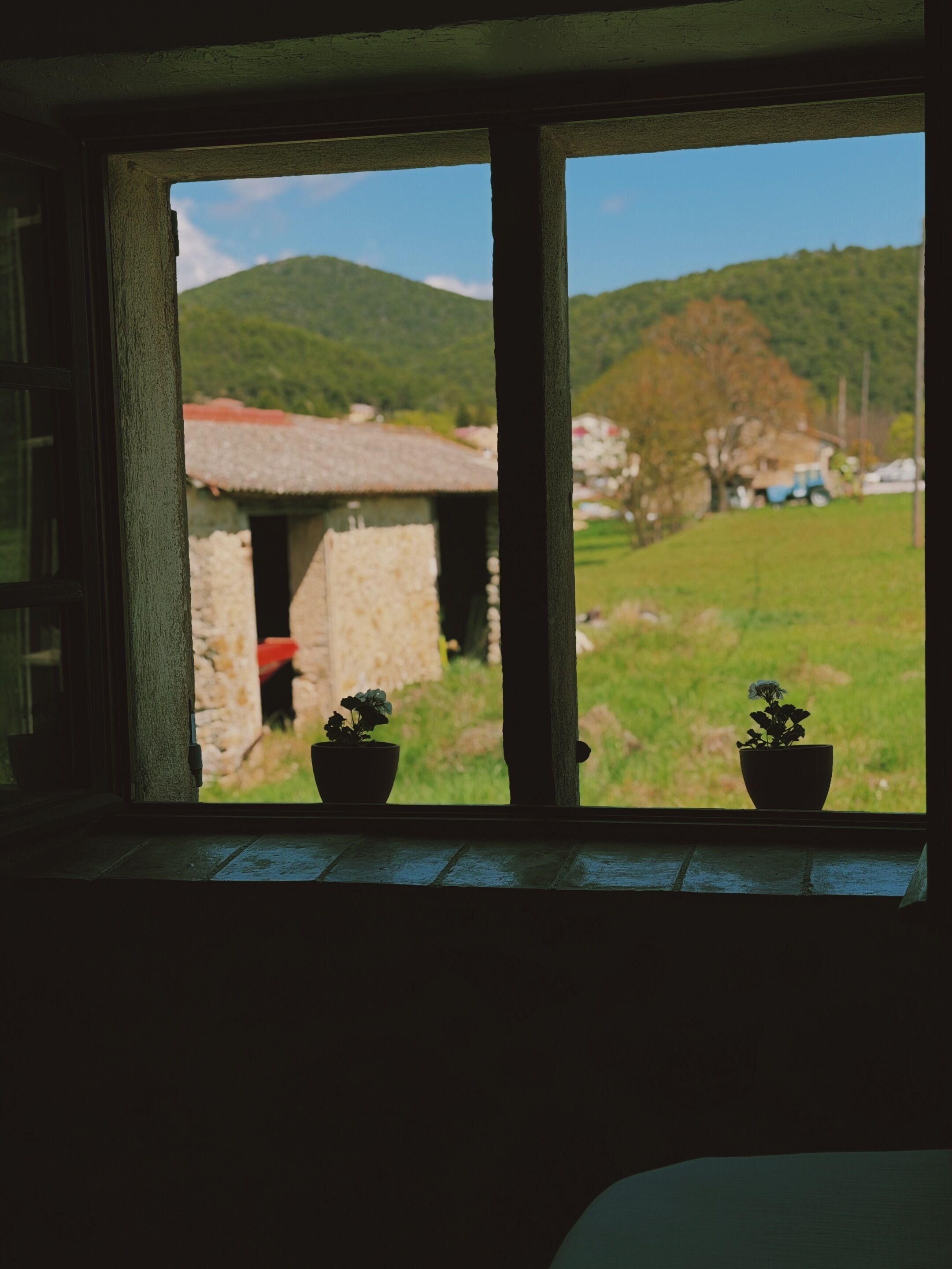 Ein Fenster mit zwei Blumentöpfen, ein Steinhaus und eine Wiese mit Hügeln im Hintergrund.