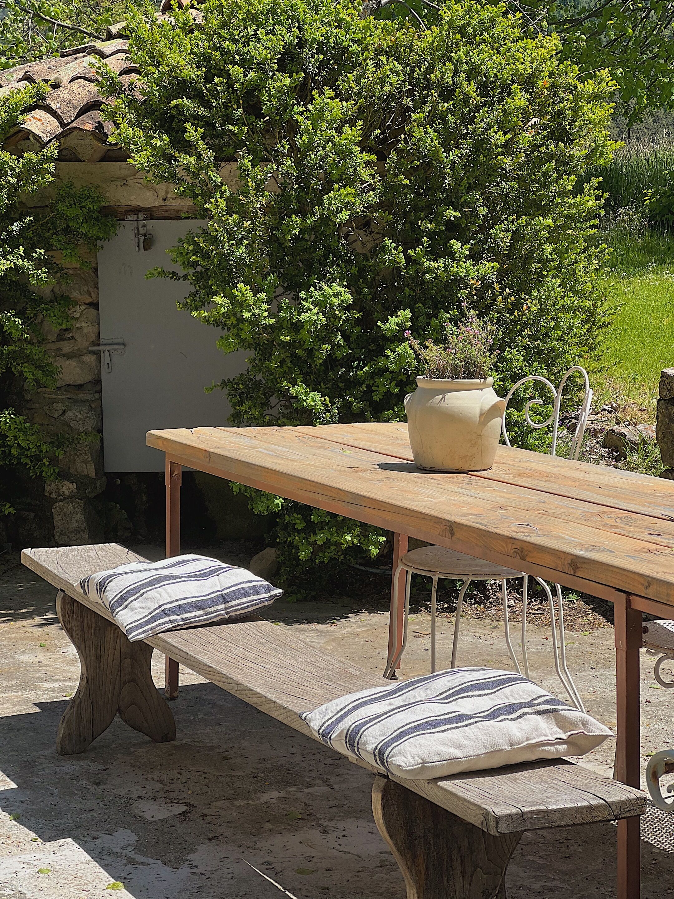 Una mesa de madera al aire libre con bancos, una planta en maceta y un muro de piedra al fondo.