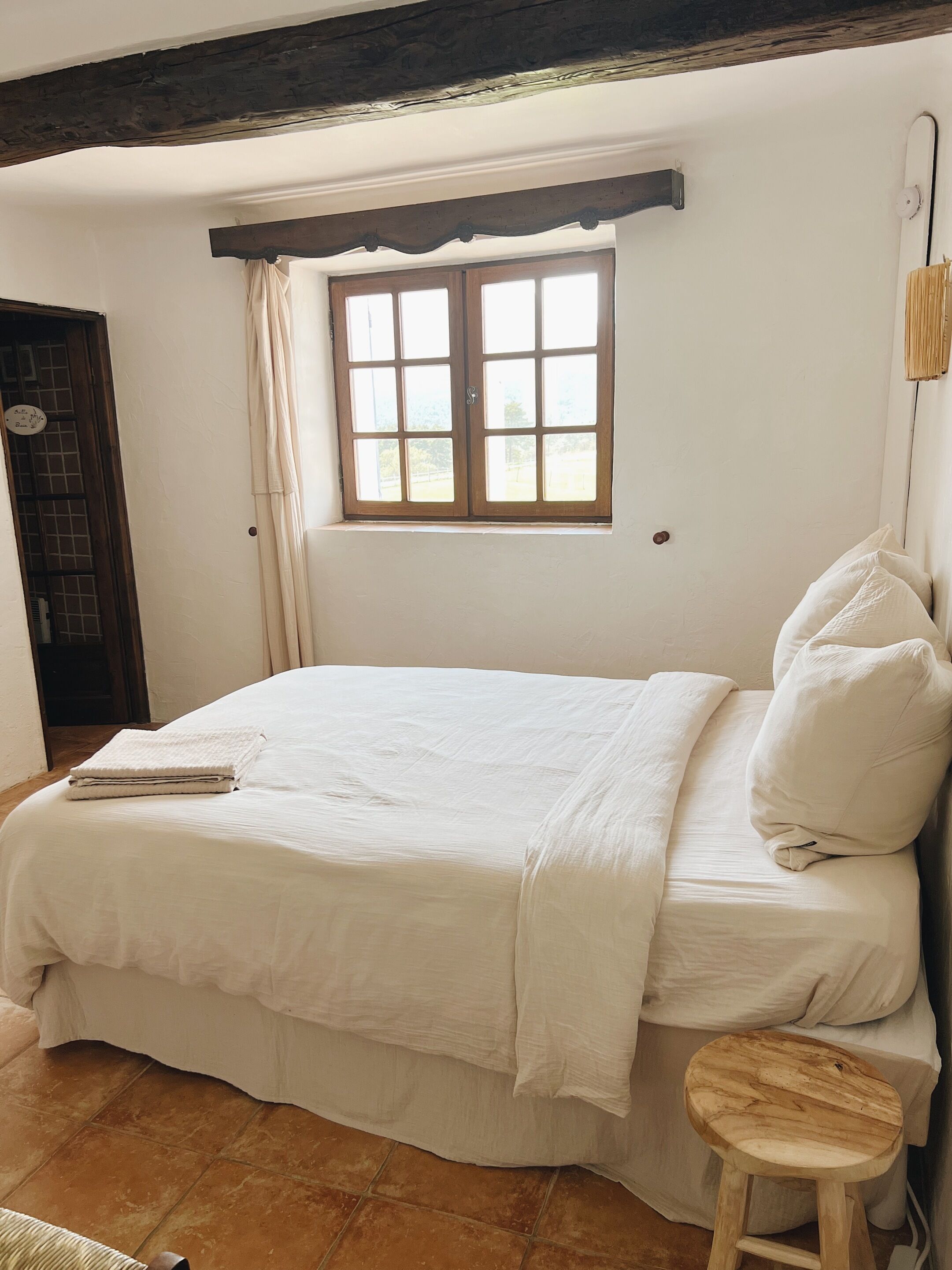 Ein Schlafzimmer mit Bett, Fenster, Tür und einem kleinen Hocker.