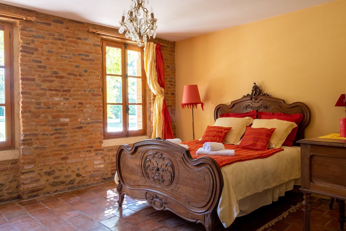 A bedroom with a brick wall, a wooden bed with red and beige bedding, a chandelier, and a window with curtains.