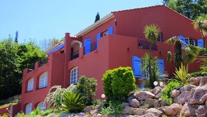 A red building with blue windows and balconies, surrounded by rocks and plants.