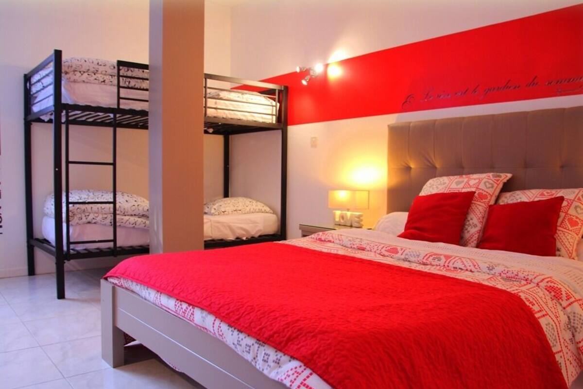 Ein Zimmer mit einem Etagenbett, einem Einzelbett mit roter Decke sowie einer Wand mit rotem Akzent und einer Leuchte.