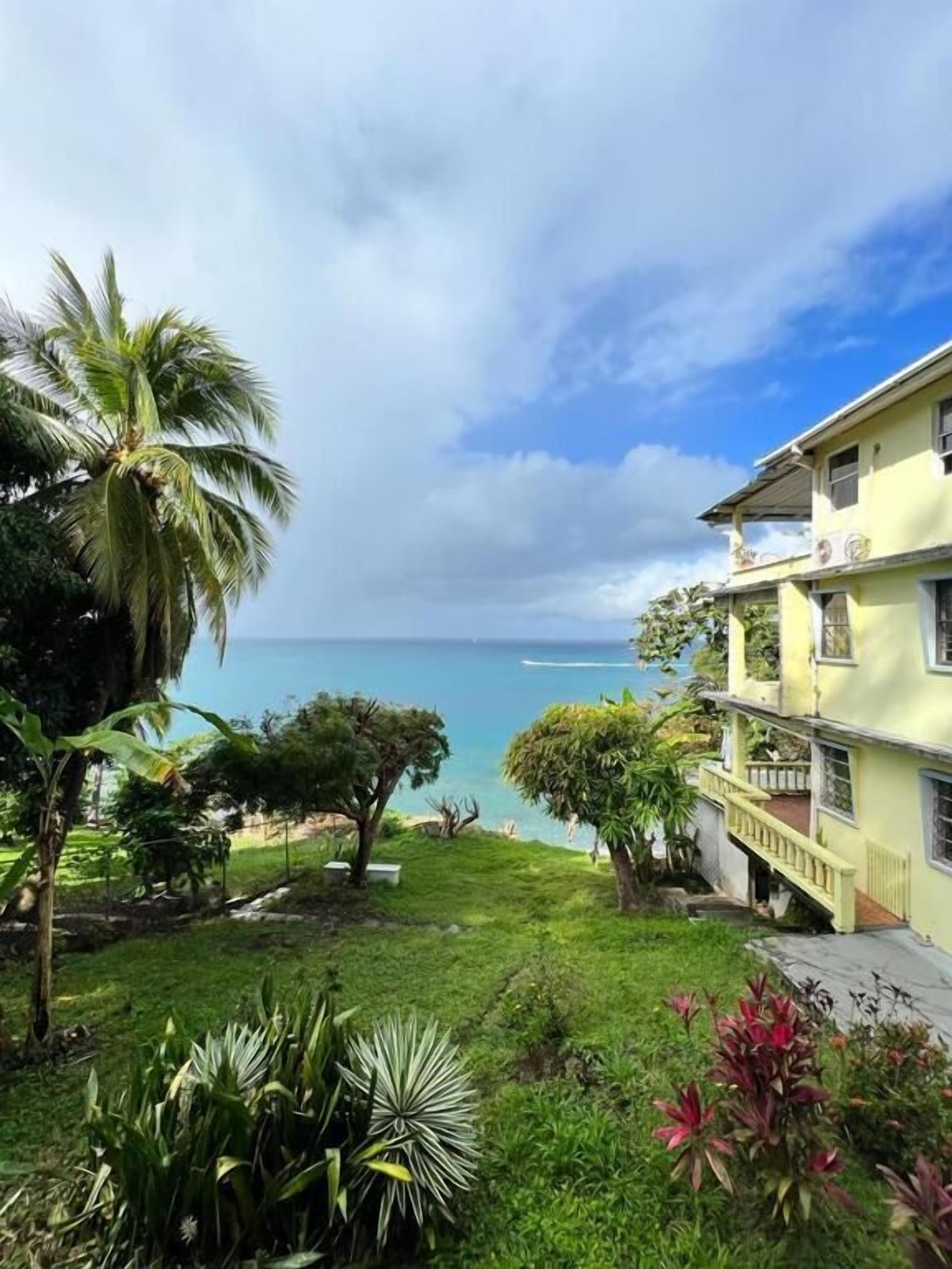 Un bâtiment situé sur la côte, doté d’un balcon donnant sur la mer, et entouré d’une végétation luxuriante et de palmiers.