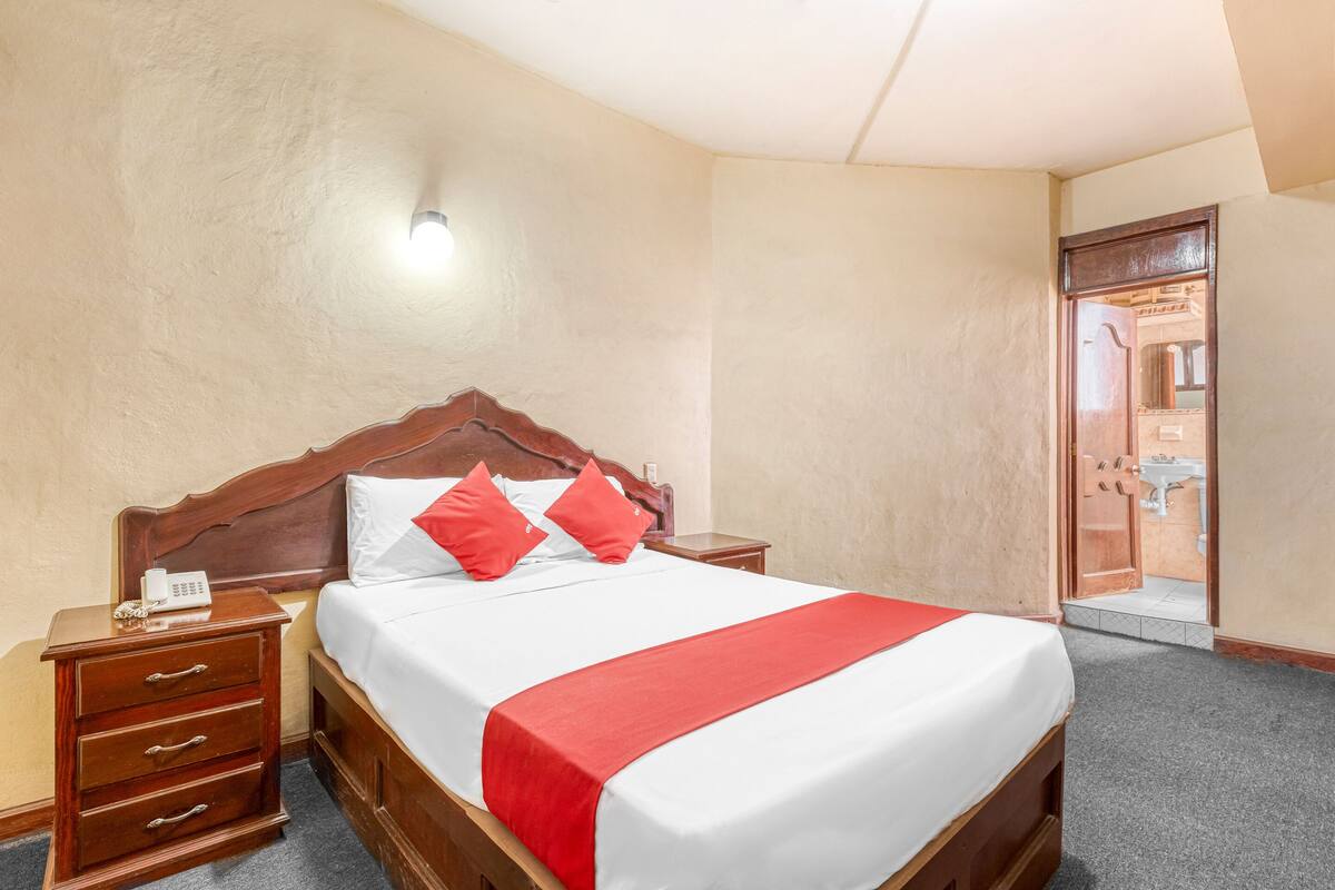 Une chambre d’hôtel avec un lit en bois, du linge de lit blanc, des oreillers rouges, une table de chevet avec un téléphone et une porte donnant sur une salle de bain.
